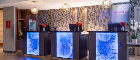 Zona de recepción de hotel con diseño moderno, que incluye un mostrador de recepción con señalización iluminada, una pantalla digital y un televisor de pared.