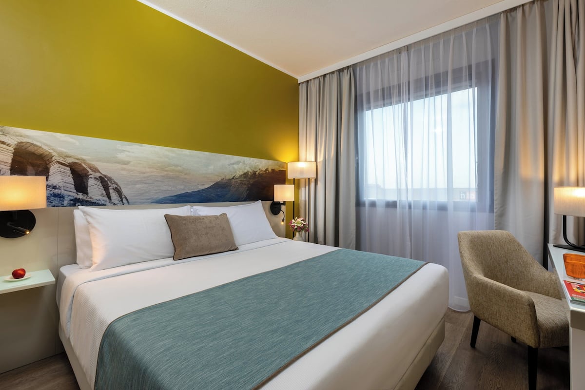 Une chambre d’hôtel comprenant un lit, deux tables de chevet, une chaise, un bureau et une fenêtre avec des rideaux.