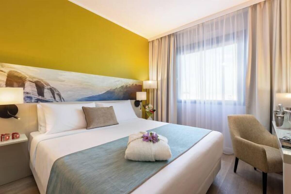 Une chambre d’hôtel avec un lit, une chaise, un bureau, un tableau au mur et une fenêtre avec des rideaux.