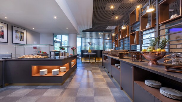A modern hotel lobby with a central reception desk, check-in counters, and a seating area. There are framed pictures on the walls, a large window providing natural light, and a decorative wall with shelves and plants. - Leonardo Hotel Verona (Verona)