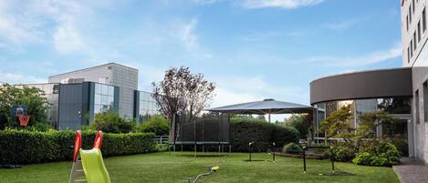 A well-maintained grassy area with playground equipment, including a slide and swings, surrounded by modern buildings and a clear sky.