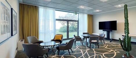 Quarto de hotel com uma janela grande, televisão de tela plana, planta de cacto e área de estar com cadeiras e uma mesa.