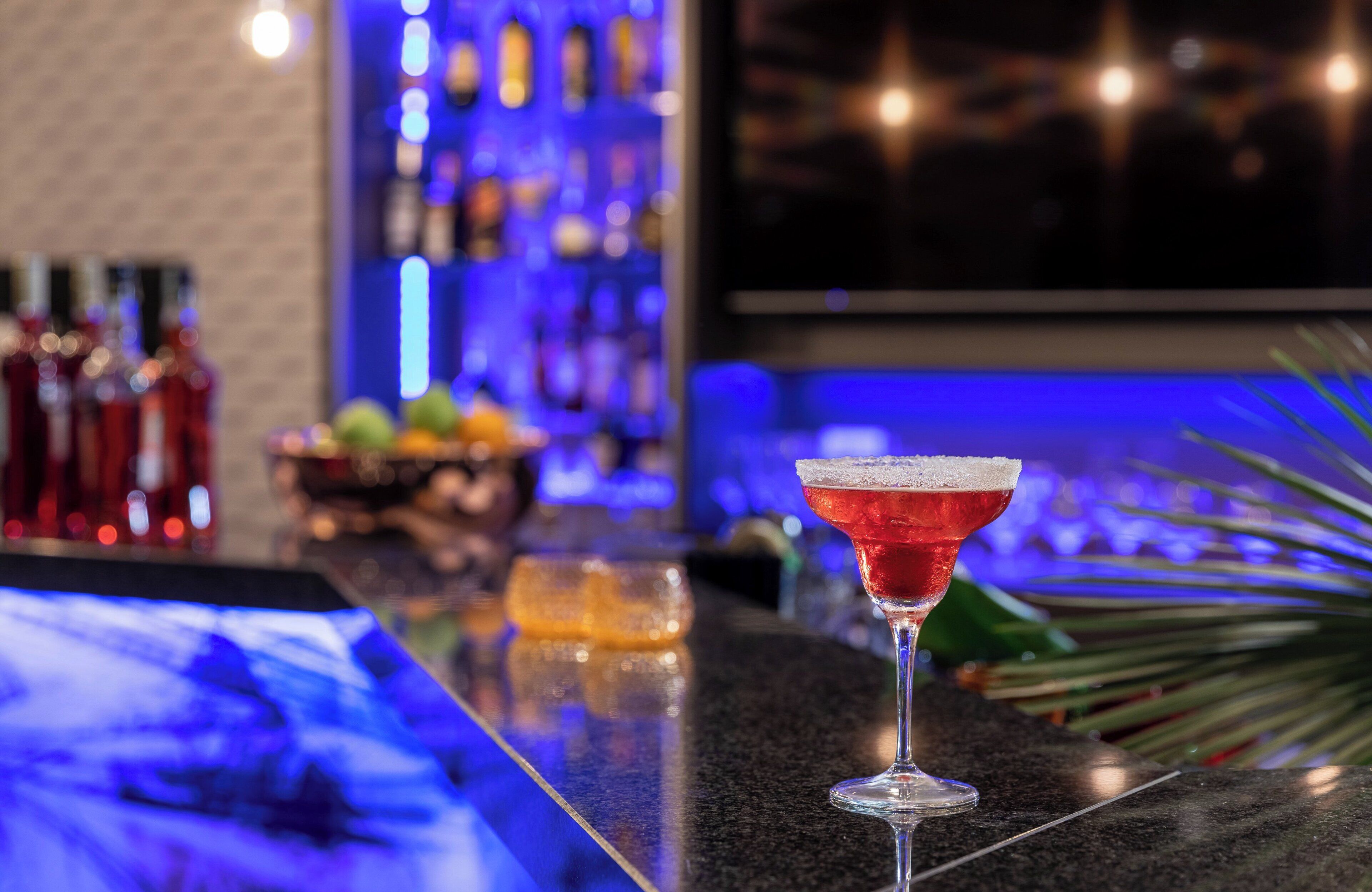 A bar with a martini glass on the counter, bottles of liquor, and a fruit bowl in the background.