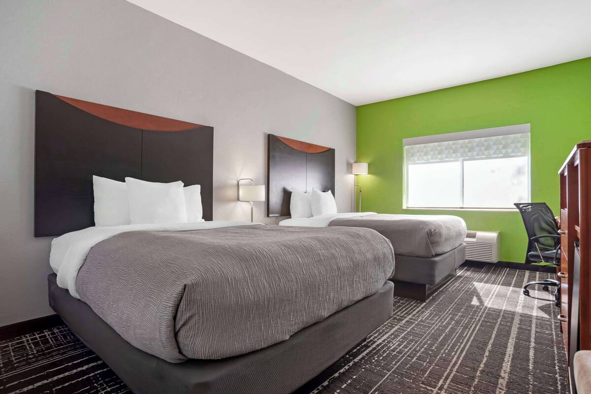 A hotel room with two beds, a large mirror, a green accent wall, a window with a geometric pattern, and a desk with a chair.