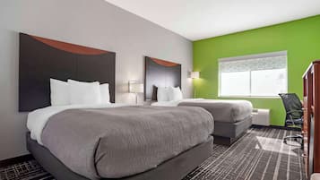 A hotel room with two beds, a large mirror, a green accent wall, a window with a geometric pattern, and a desk with a chair.