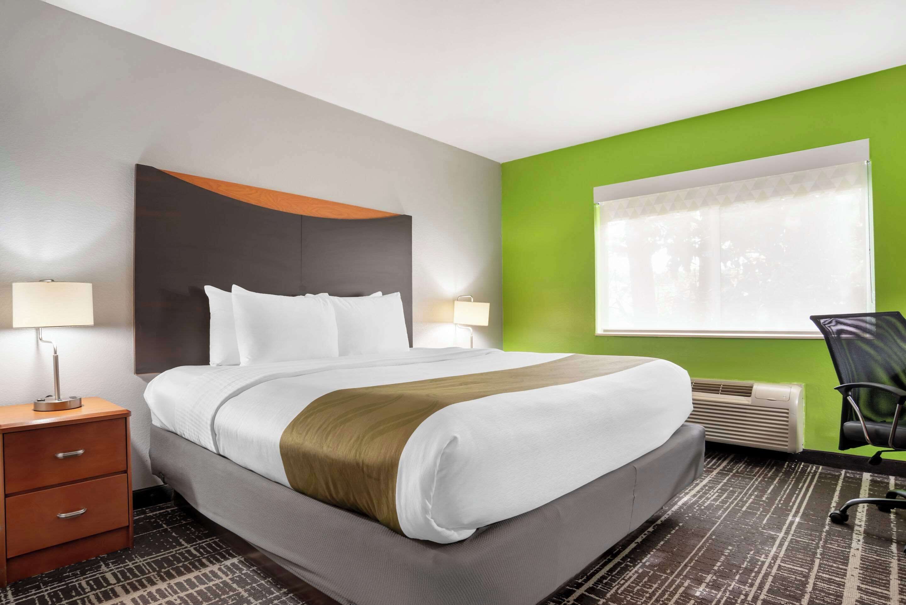 A hotel room with a large bed, a nightstand, a lamp, a chair, and a window with blinds.