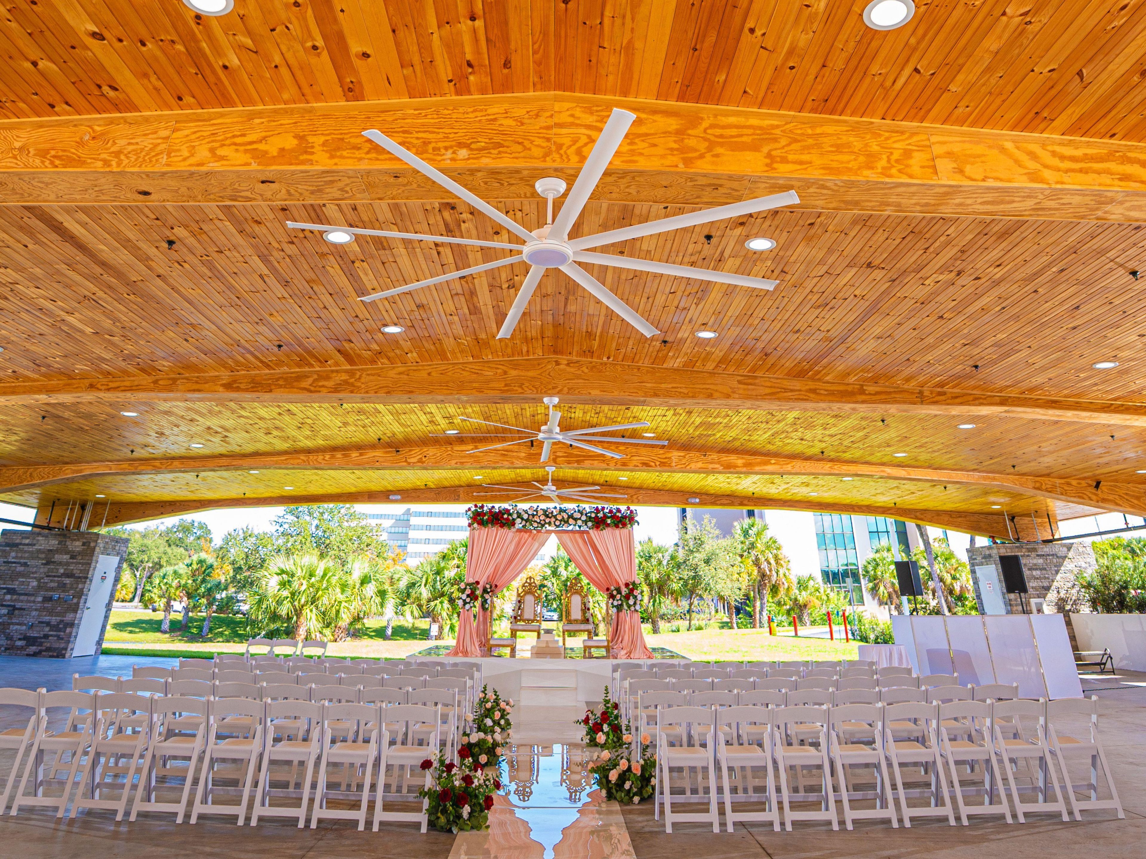 An indoor venue with a wooden ceiling and multiple ceiling fans, arranged seating, and a stage with floral decorations.