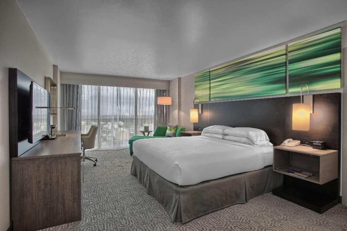 A hotel room with a bed, desk, chair, and a large window with curtains.
