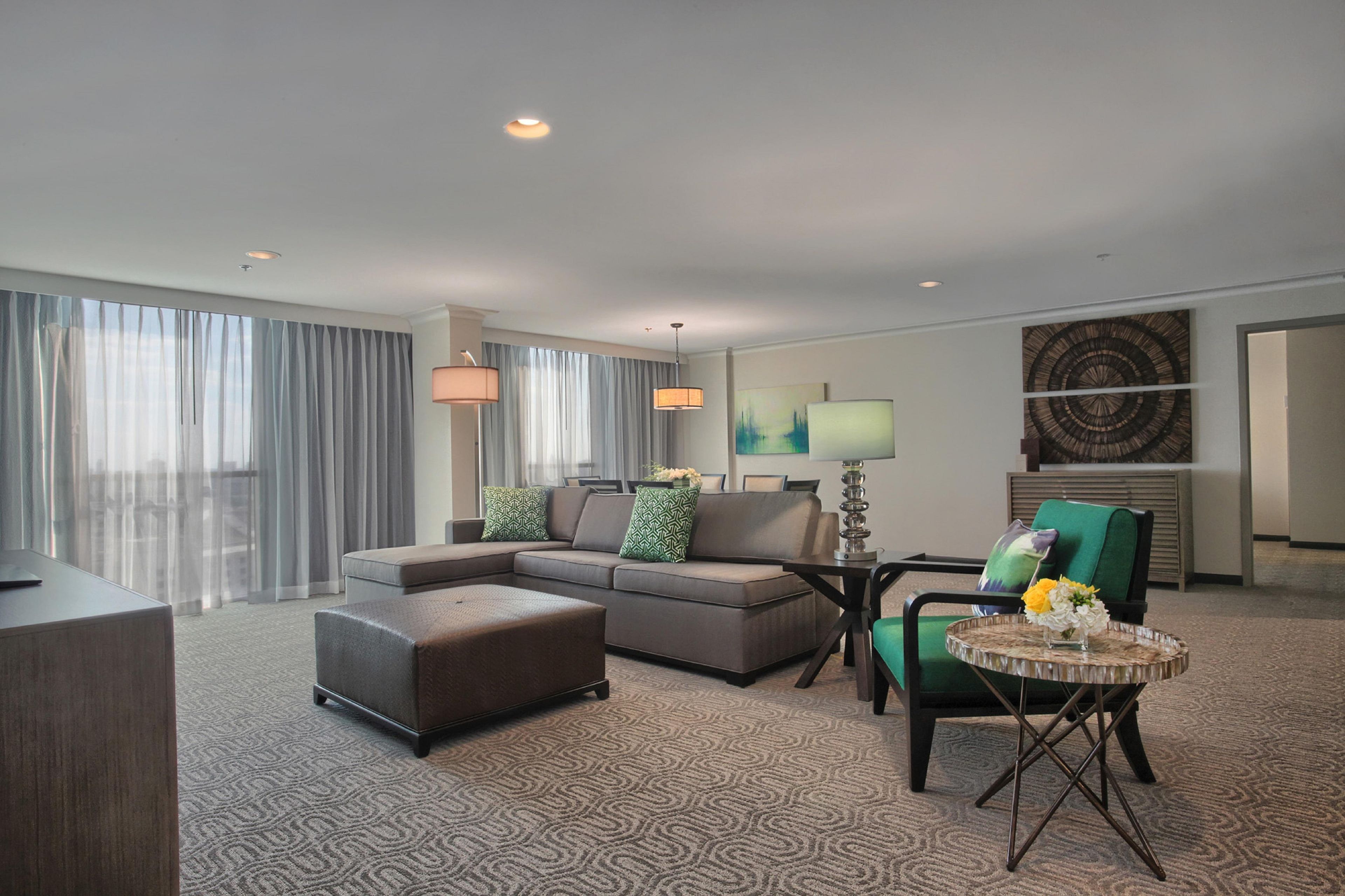 Une chambre d’hôtel moderne avec un canapé d’angle, un petit pouf, un fauteuil vert, une table basse ronde et une grande fenêtre avec des rideaux légers.