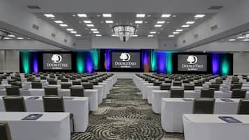 Un salón de conferencias con filas de mesas y sillas, un escenario con una pantalla que muestra el logotipo de DoubleTree by Hilton, y una alfombra estampada.