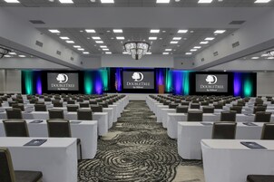 Un salón de conferencias con filas de mesas y sillas, un escenario con una pantalla que muestra el logotipo de DoubleTree by Hilton, y una alfombra estampada.