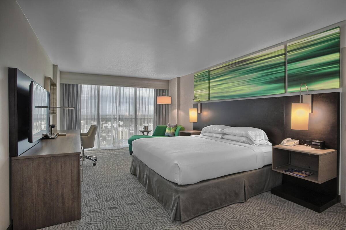 A hotel room with a bed, desk, chair, and a large window with curtains.