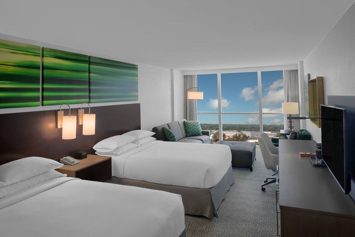 A hotel room with two beds, a desk, a TV, and a large window offering a city view.
