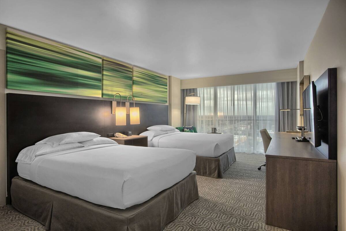 A hotel room with two beds, a desk, a TV, and a large window with curtains.