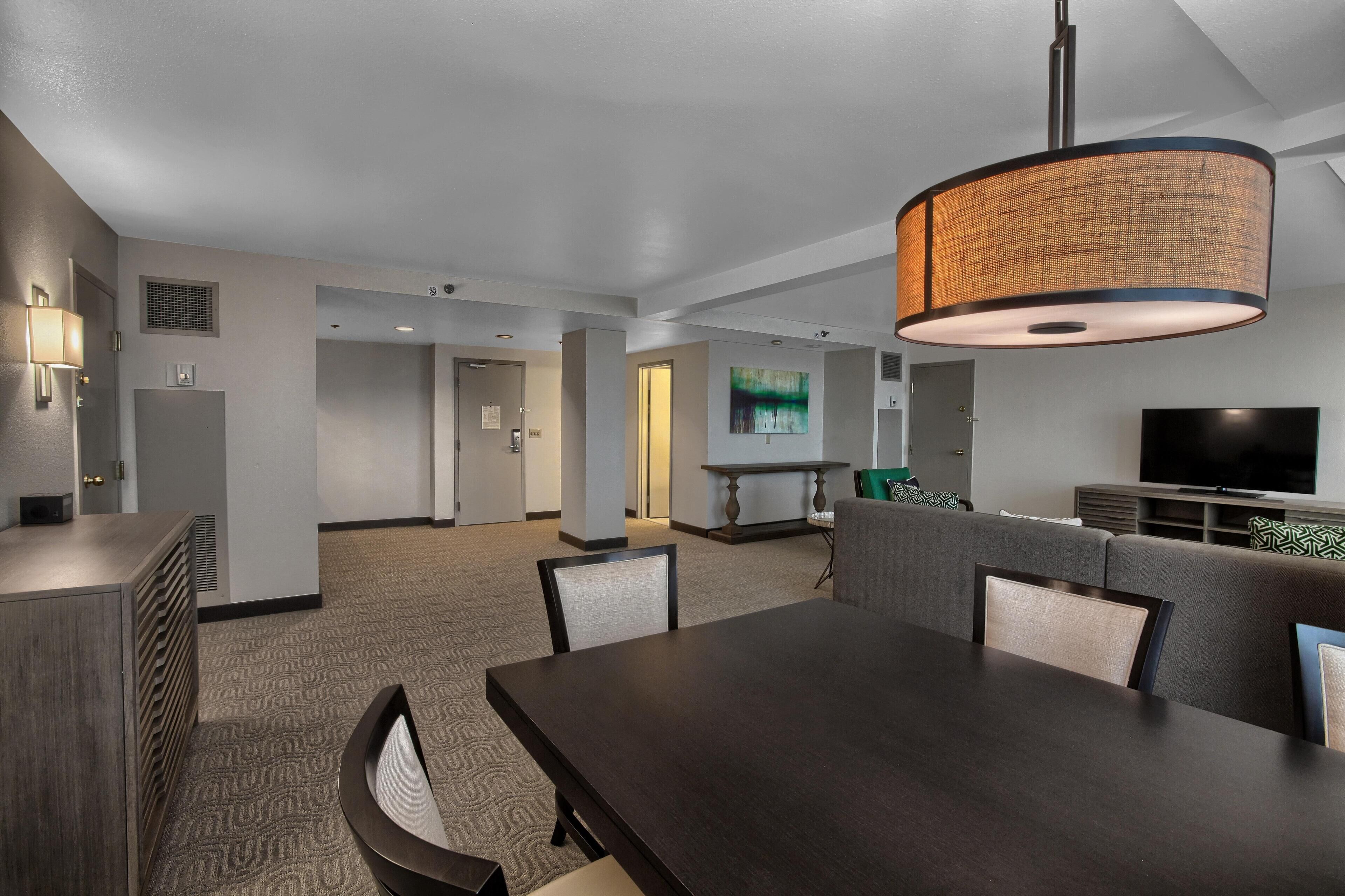 A modern hotel room with a dining area, a sofa, a TV, and a large window.