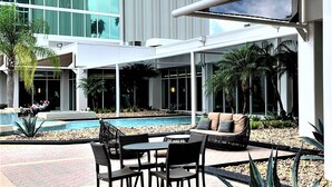 An outdoor pool area with a covered seating area, surrounded by palm trees and a modern building in the background. - DoubleTree by Hilton at the Entrance to Universal Orlando (Orlando)