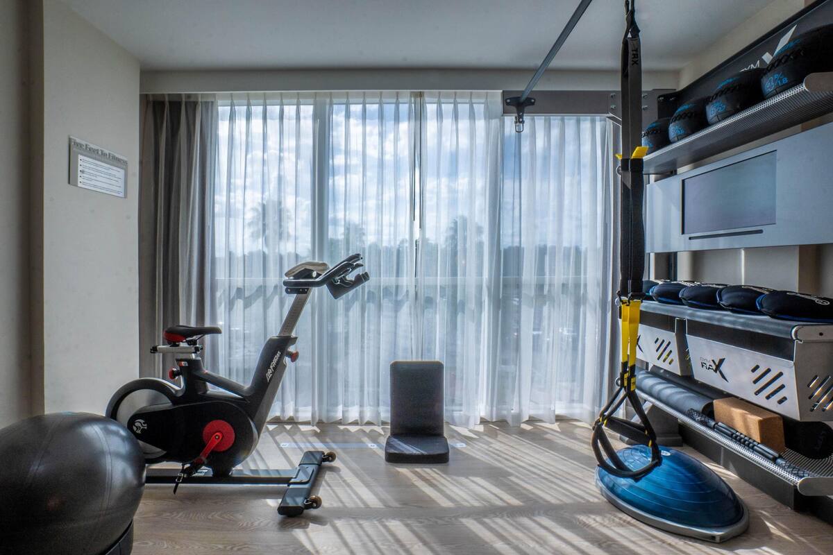 A room with a stationary bike, a rowing machine, a blue exercise mat, and a rack with bags.
