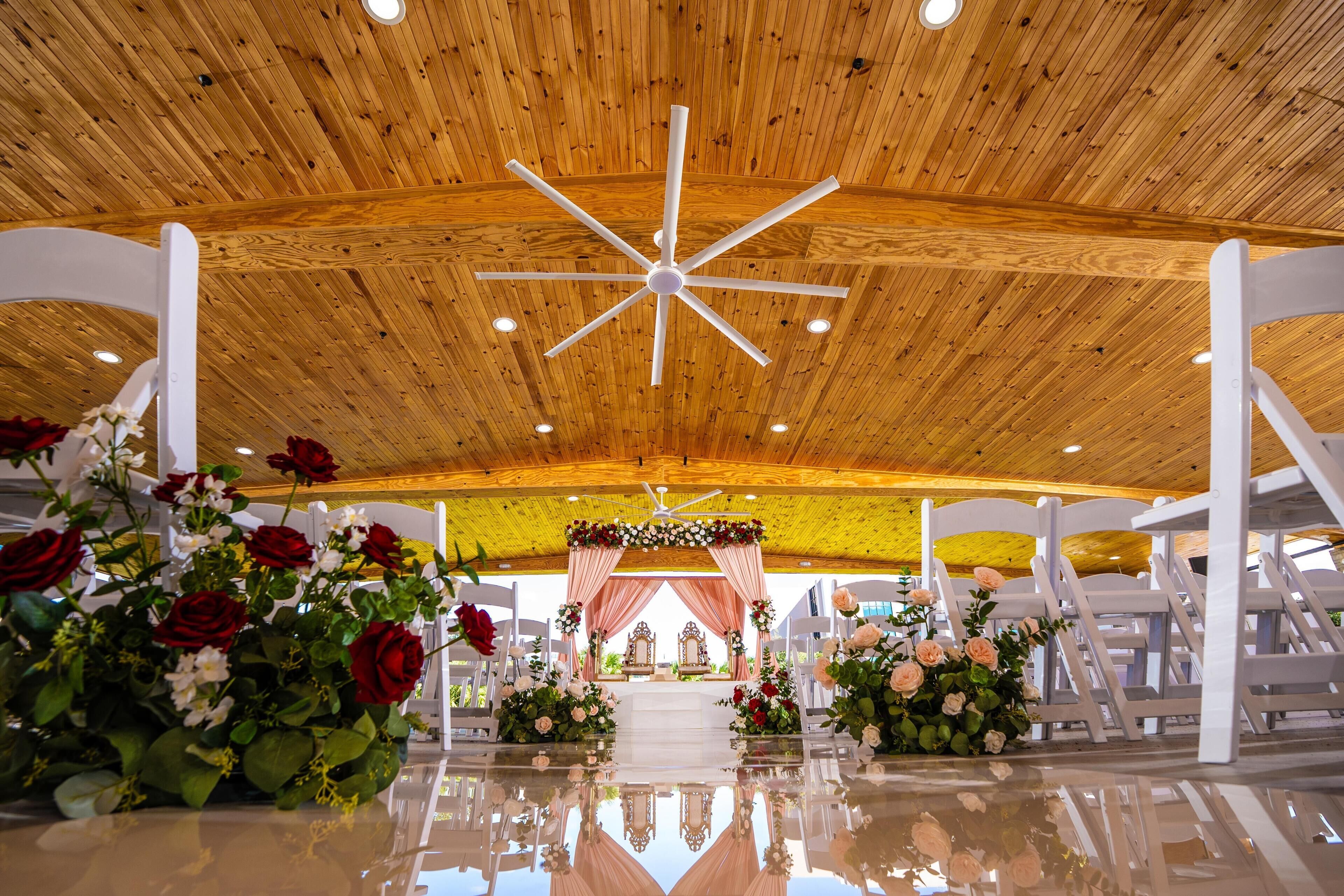 A wooden ceiling with a central fan, white chairs arranged in rows, and floral decorations at the front.