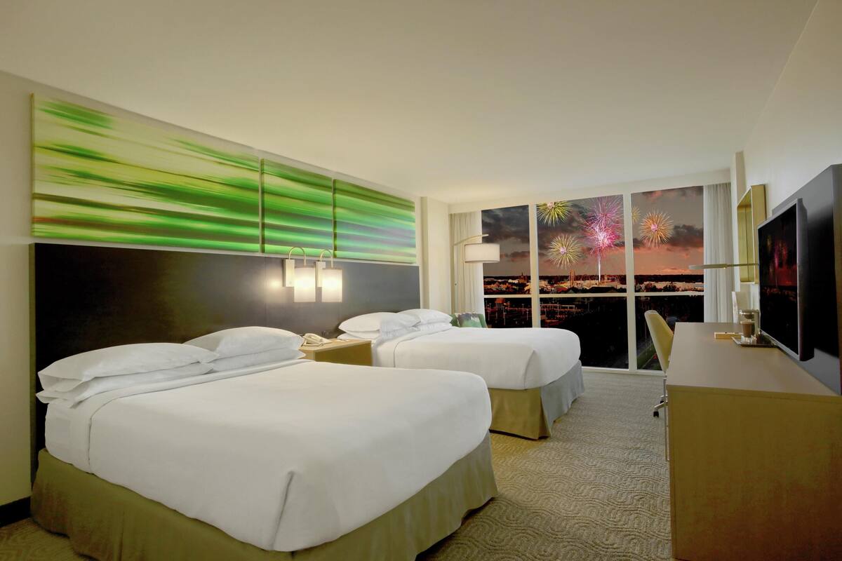 A hotel room with two beds, a desk, a television, and a large window with a city view.