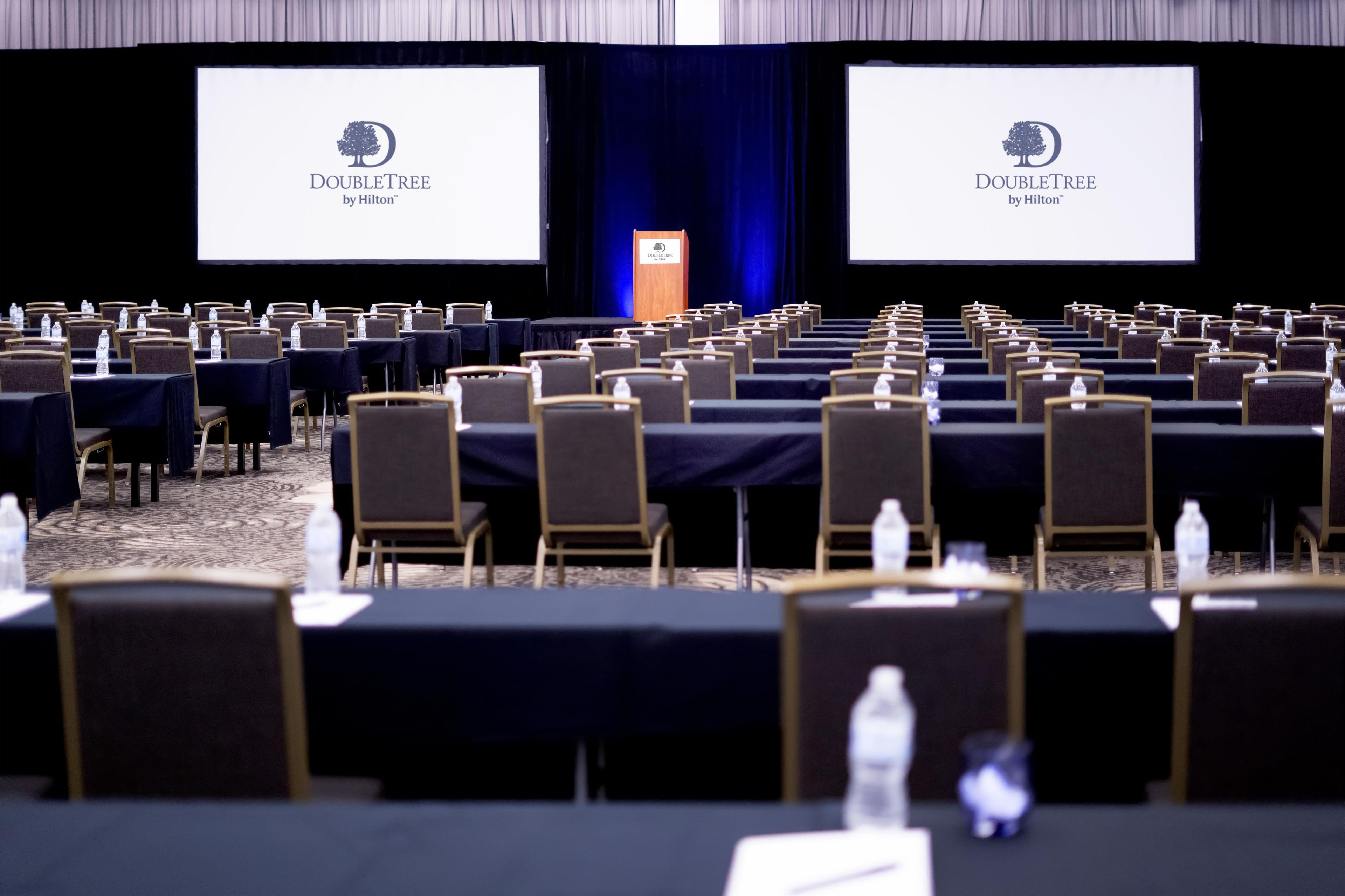 A conference room with rows of tables and chairs, two large screens displaying the DoubleTree by Hilton logo, and a podium.