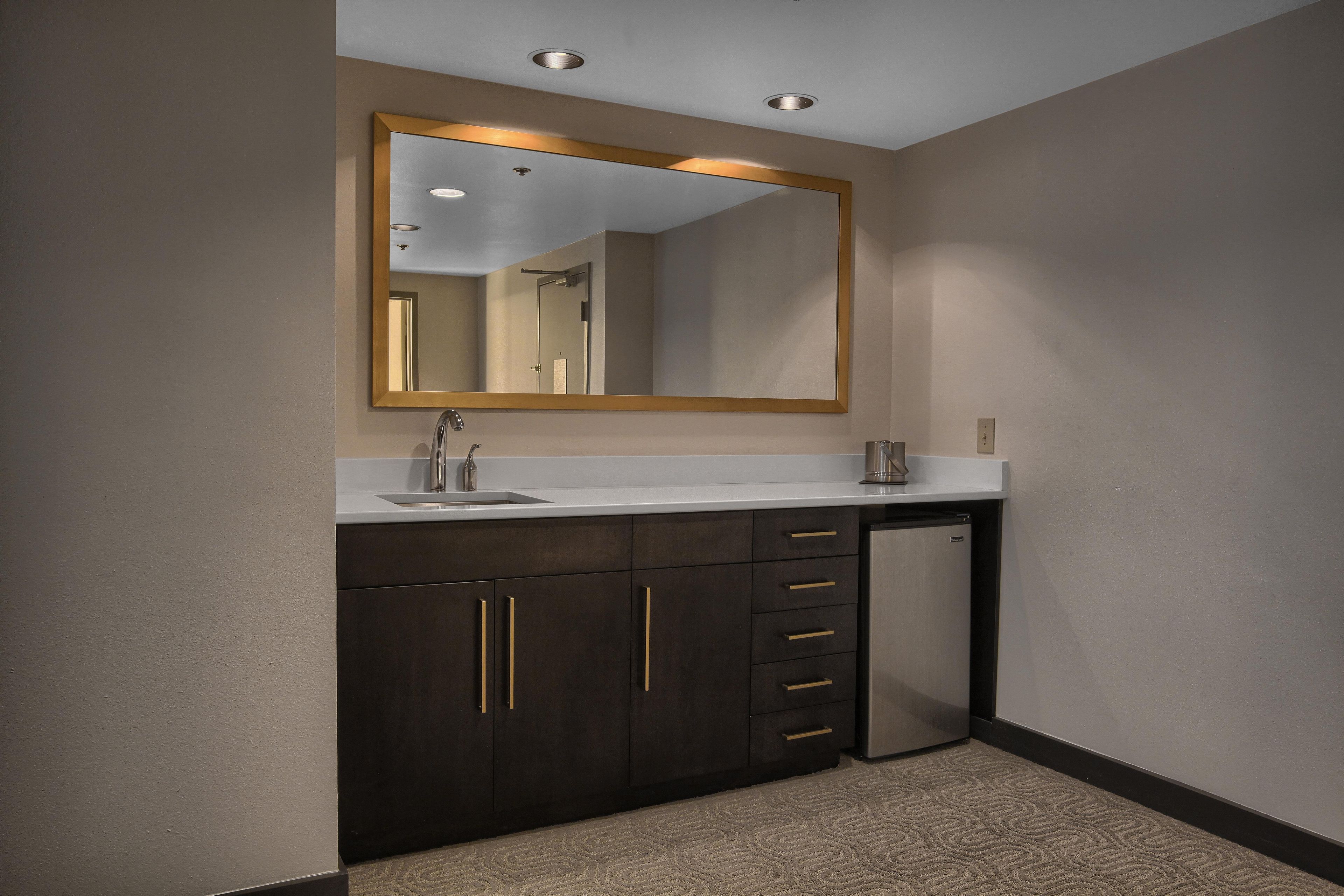 A modern bathroom with a large mirror, dark cabinets, a sink, and a compact refrigerator.