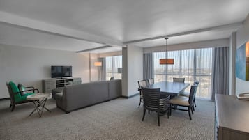 Une chambre d’hôtel avec un canapé, une table à manger avec des chaises, une télévision et une grande fenêtre avec des rideaux.