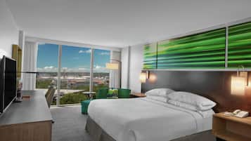 A hotel room with a large bed, a desk, a chair, a TV, a phone, and a view of the city.