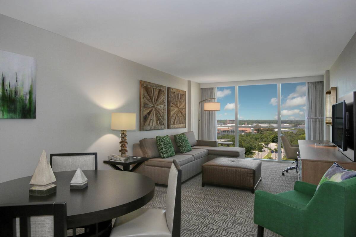A modern hotel room with a dining area, a sofa, a green armchair, a television, and large windows offering a city view.