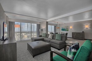 A modern hotel room with a sectional sofa, a small ottoman, a glass coffee table, and a flat-screen TV.