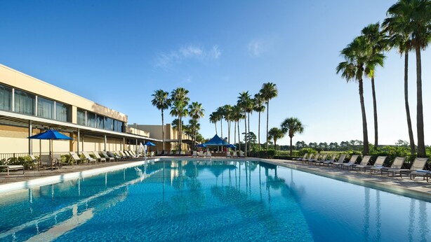 A swimming pool with lounge chairs and palm trees, surrounded by a building with large windows. - DoubleTree by Hilton at the Entrance to Universal Orlando (Orlando)