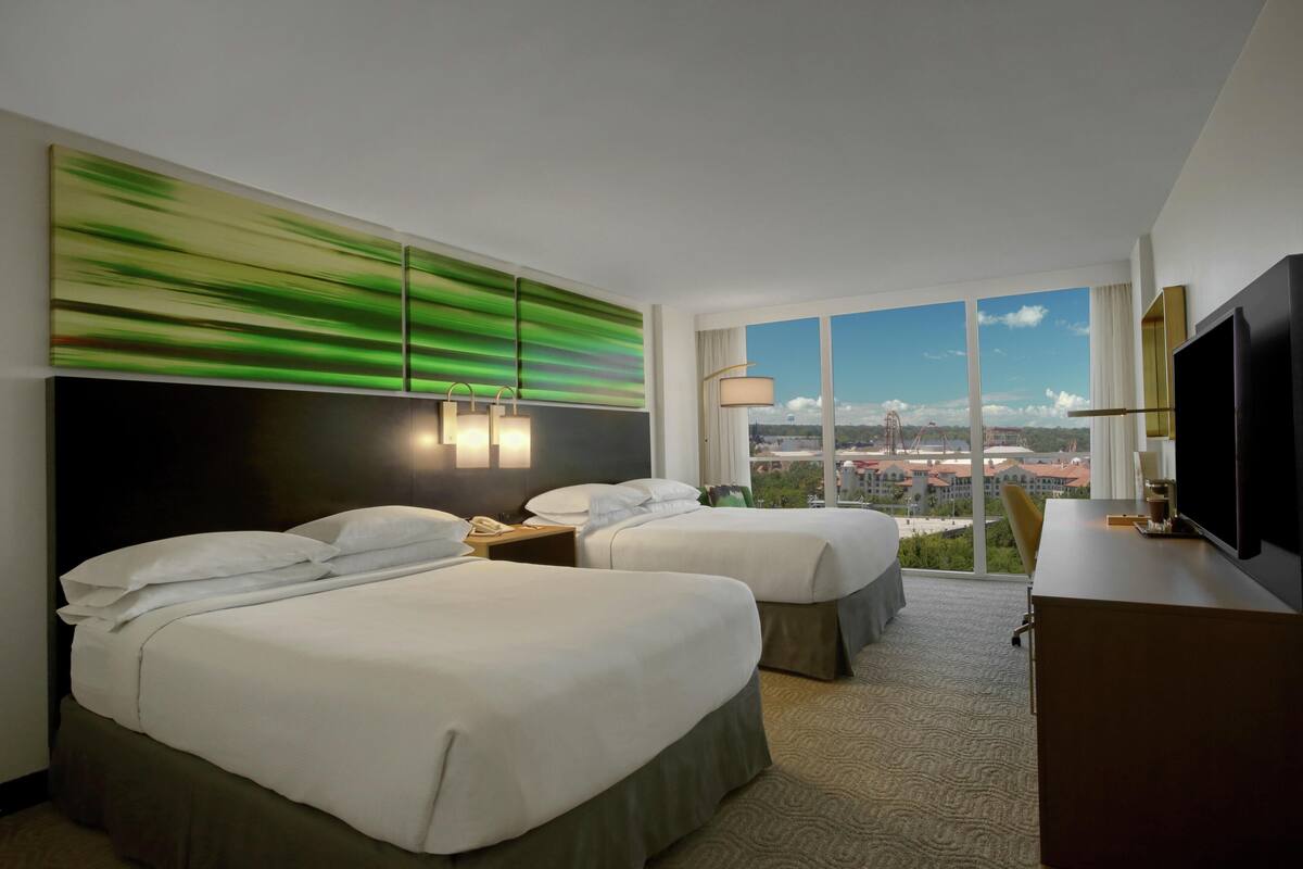 A hotel room with two beds, a large window with a city view, a flat-screen TV, and a modern abstract painting on the wall.