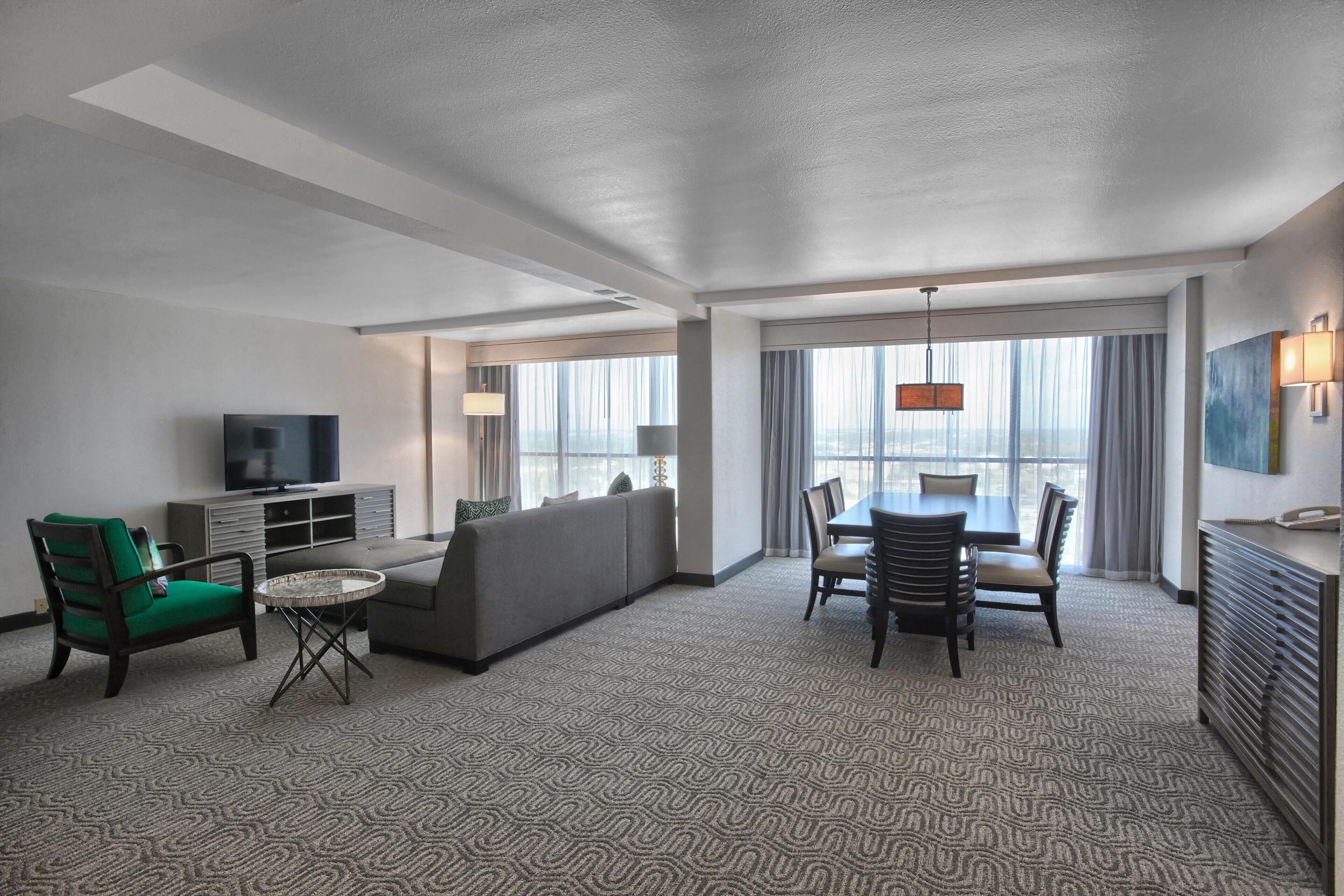 A hotel room with a sofa, chairs, a dining table, a TV, and a large window.