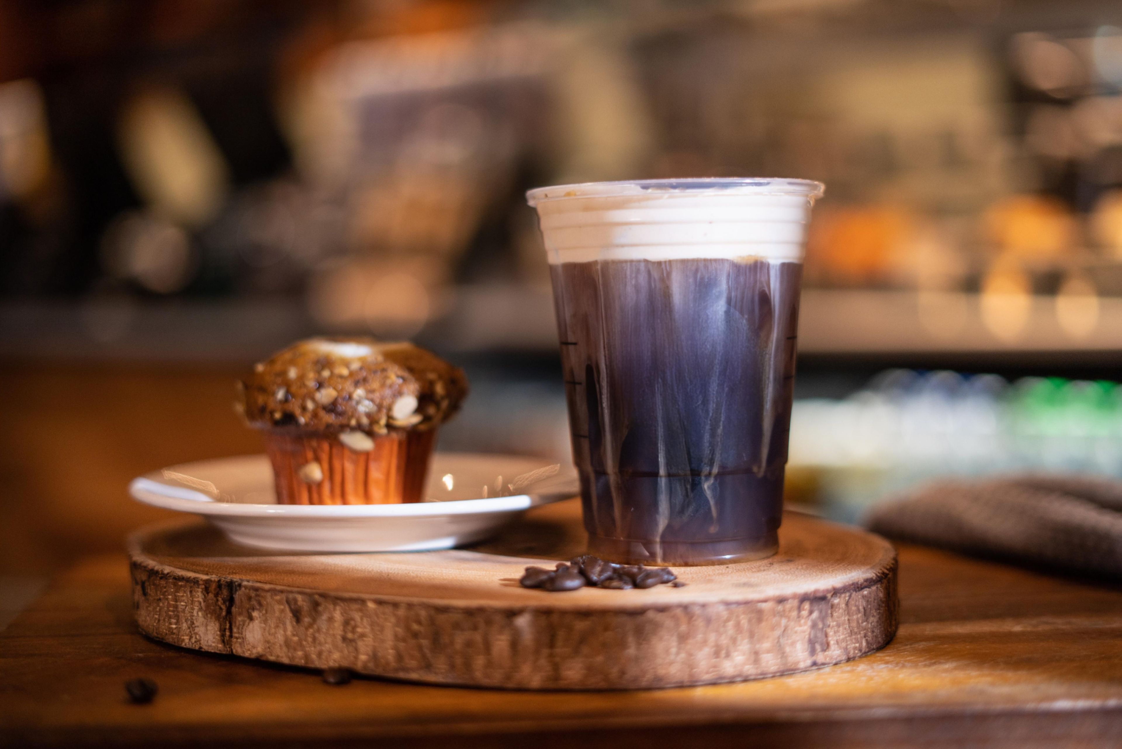 Ein Eiskaffee in einer Tasse auf einem Holzuntersetzer, daneben ein Muffin auf einem kleinen Teller.