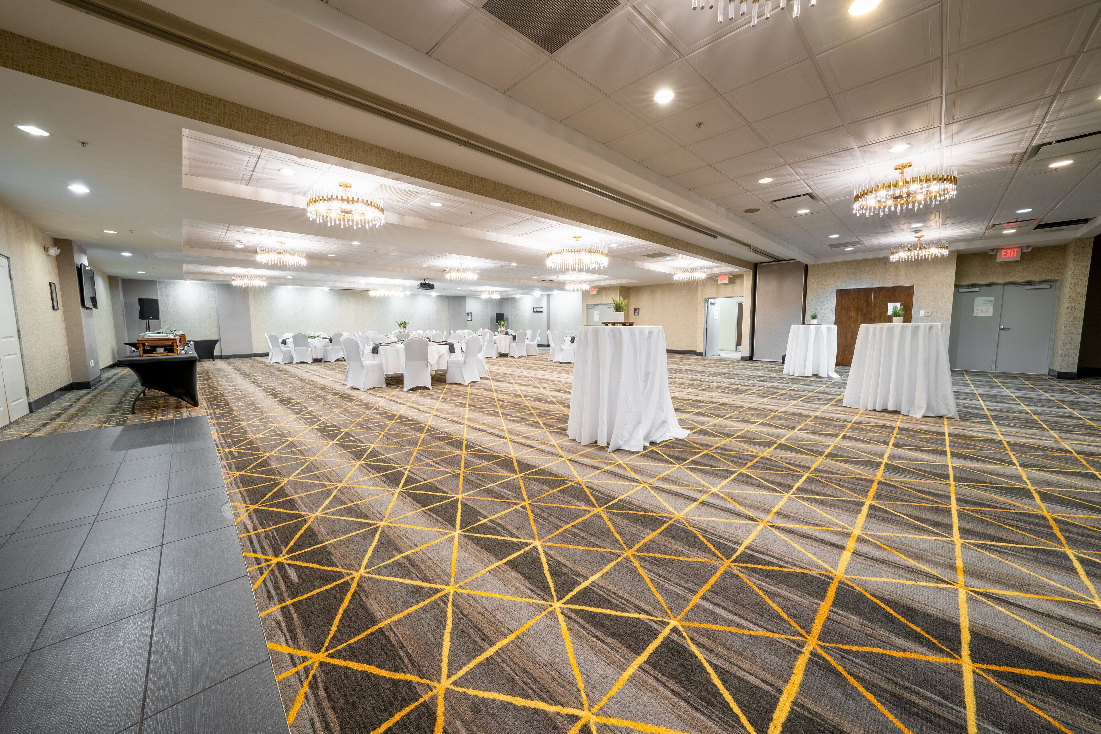A spacious conference room with a patterned carpet, multiple tables with white tablecloths, and chandeliers on the ceiling.