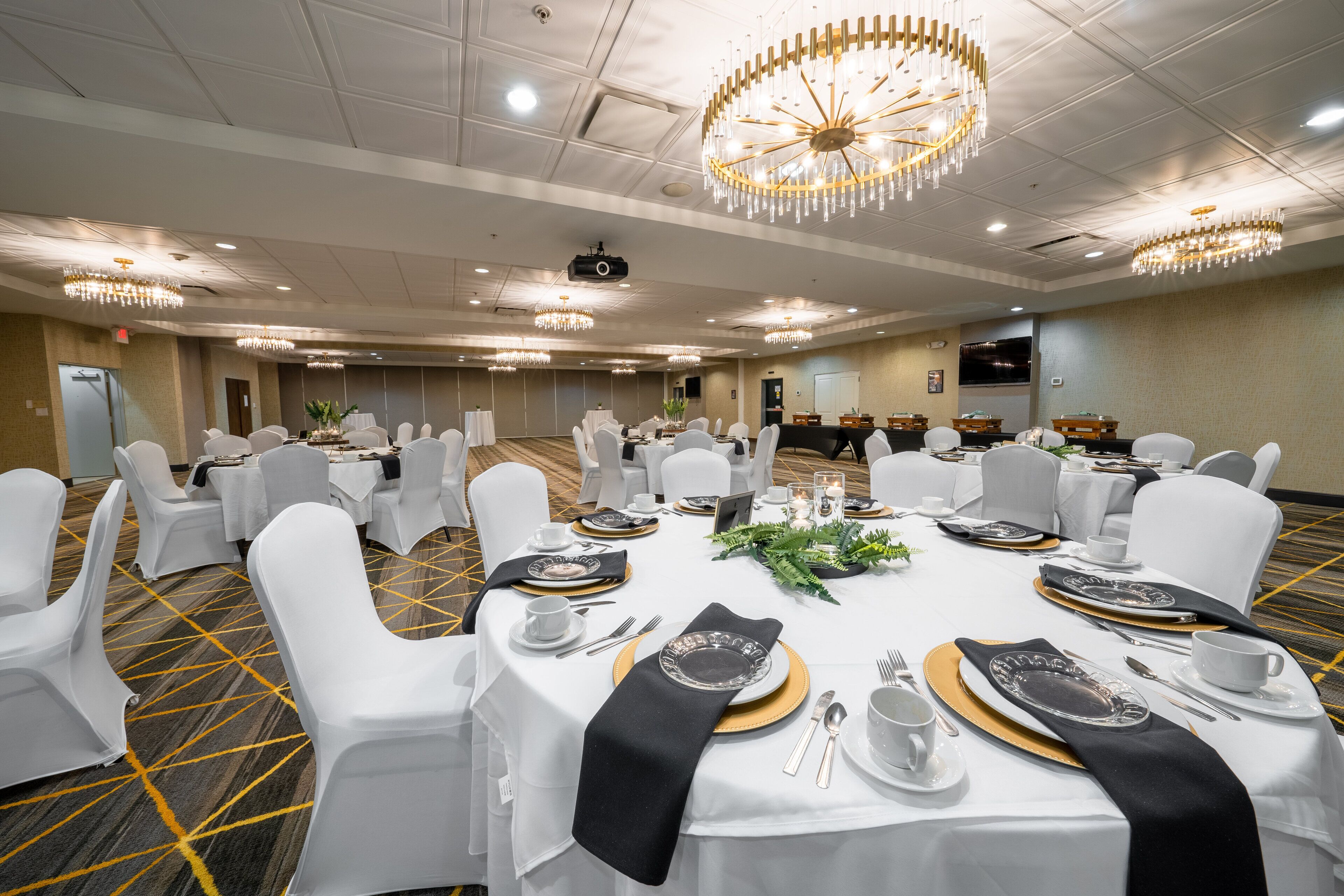 Una sala da banchetto con tavoli rotondi apparecchiati per un evento formale, con sedie bianche, piatti unici dorati e un lampadario.