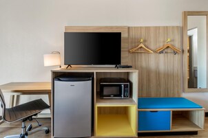 Habitación de hotel con escritorio, televisión de pantalla plana, refrigerador, microondas, una silla y armario con perchas.