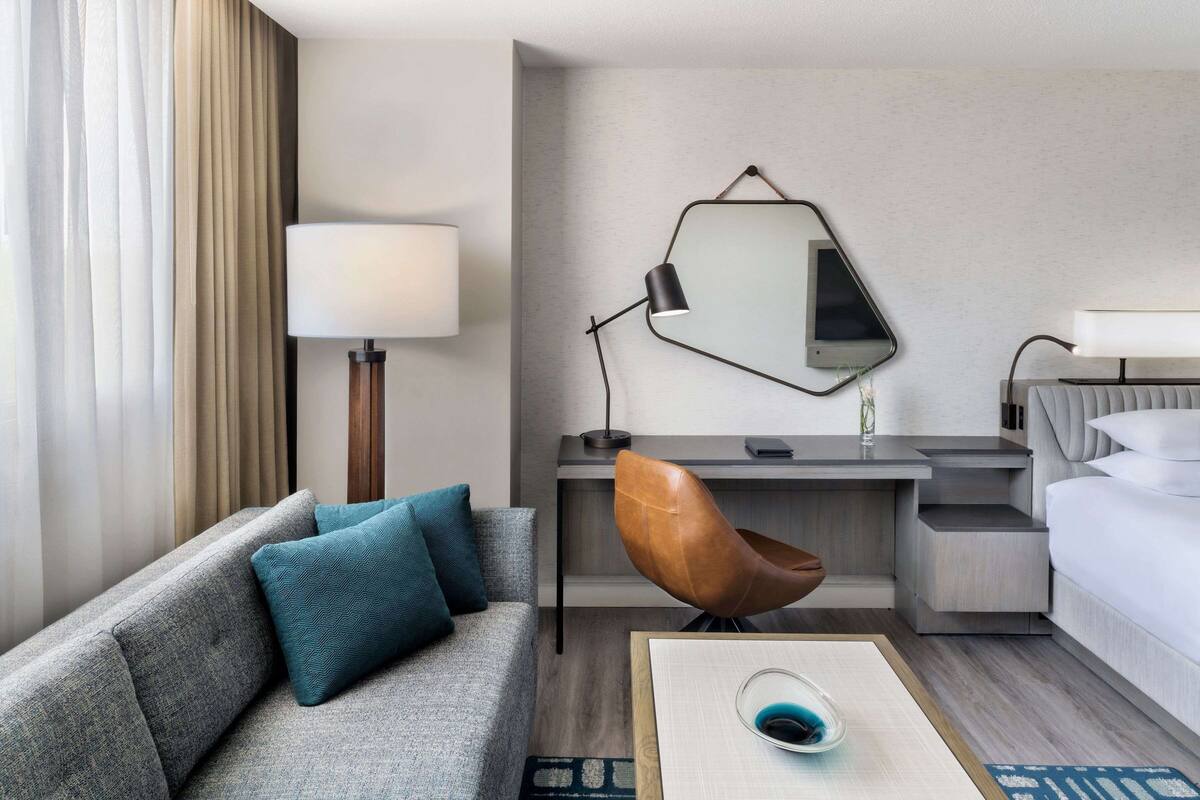 A modern hotel room with a grey sofa, teal pillows, a wooden coffee table, a desk with a laptop, a brown chair, a bed with white linens, and a large mirror on the wall.