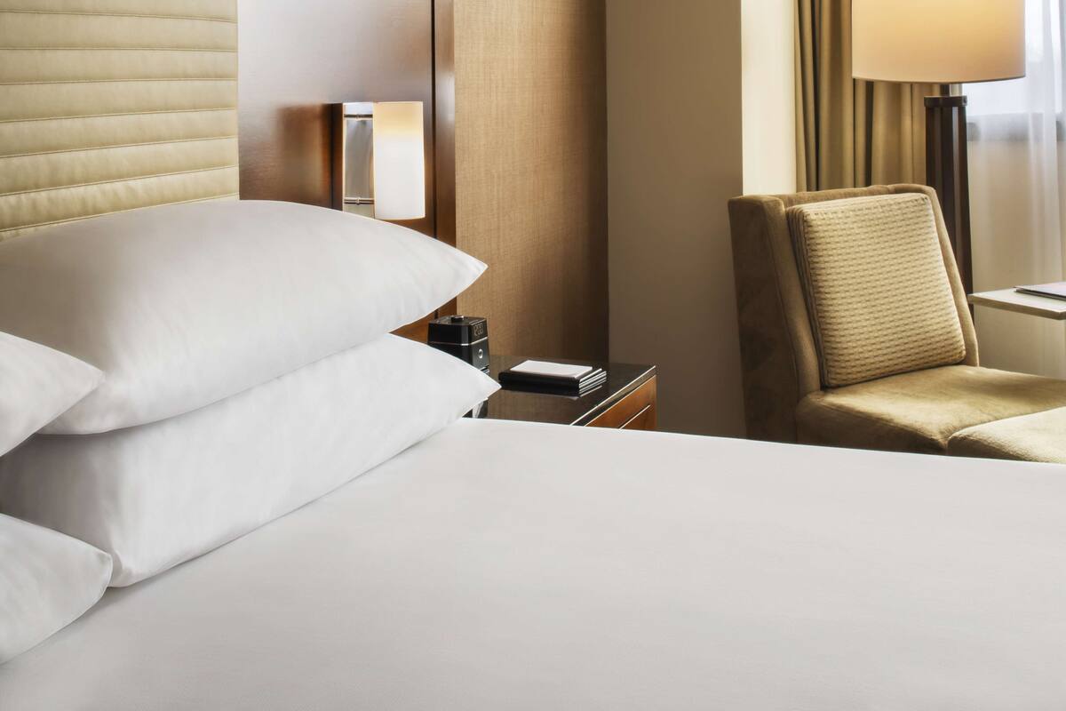 A hotel room with a neatly made bed, a bedside table with a lamp, a chair, and a window with curtains.