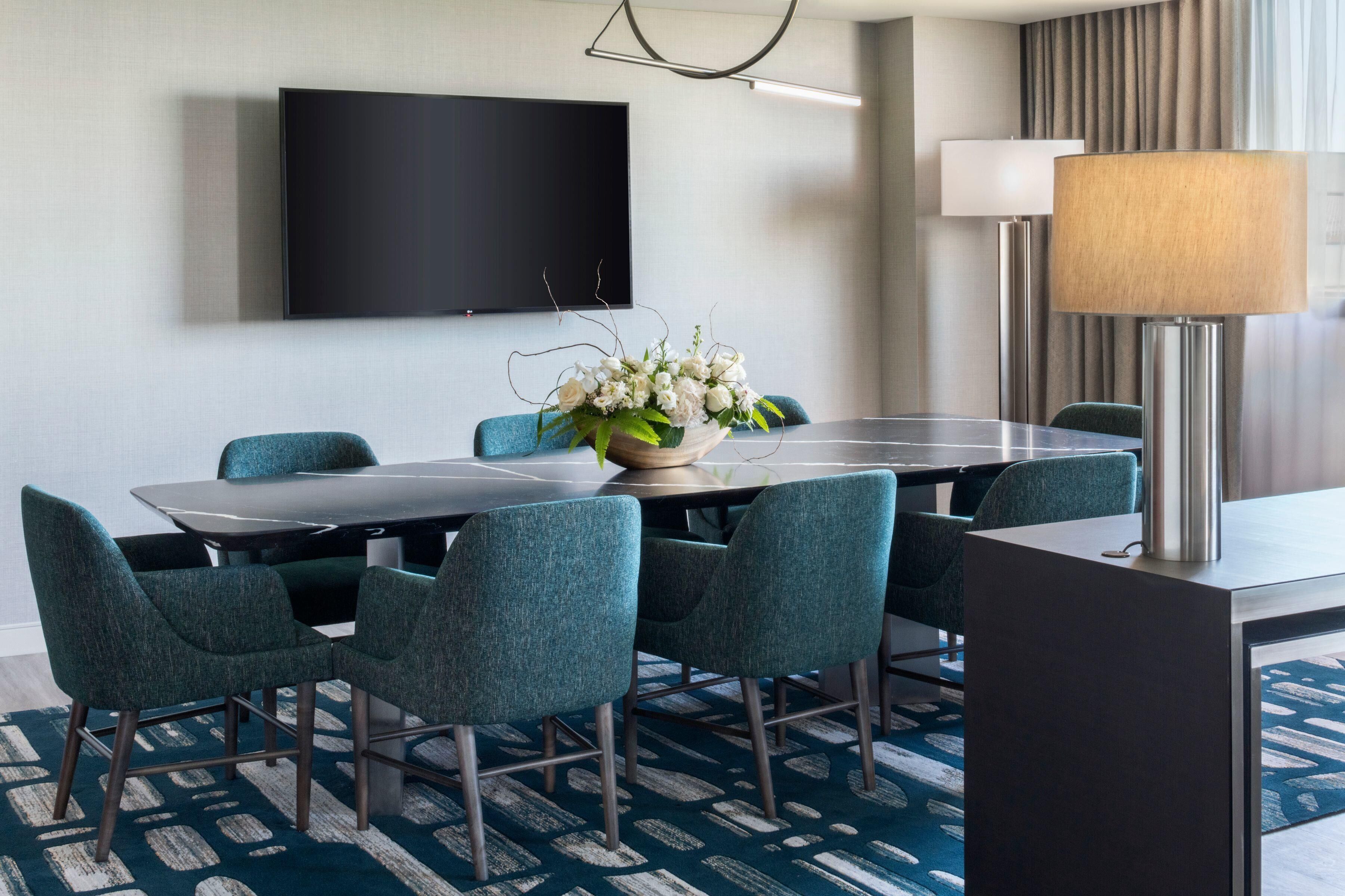A conference room with a large table, chairs, a flat-screen TV, a flower arrangement, and a lamp.