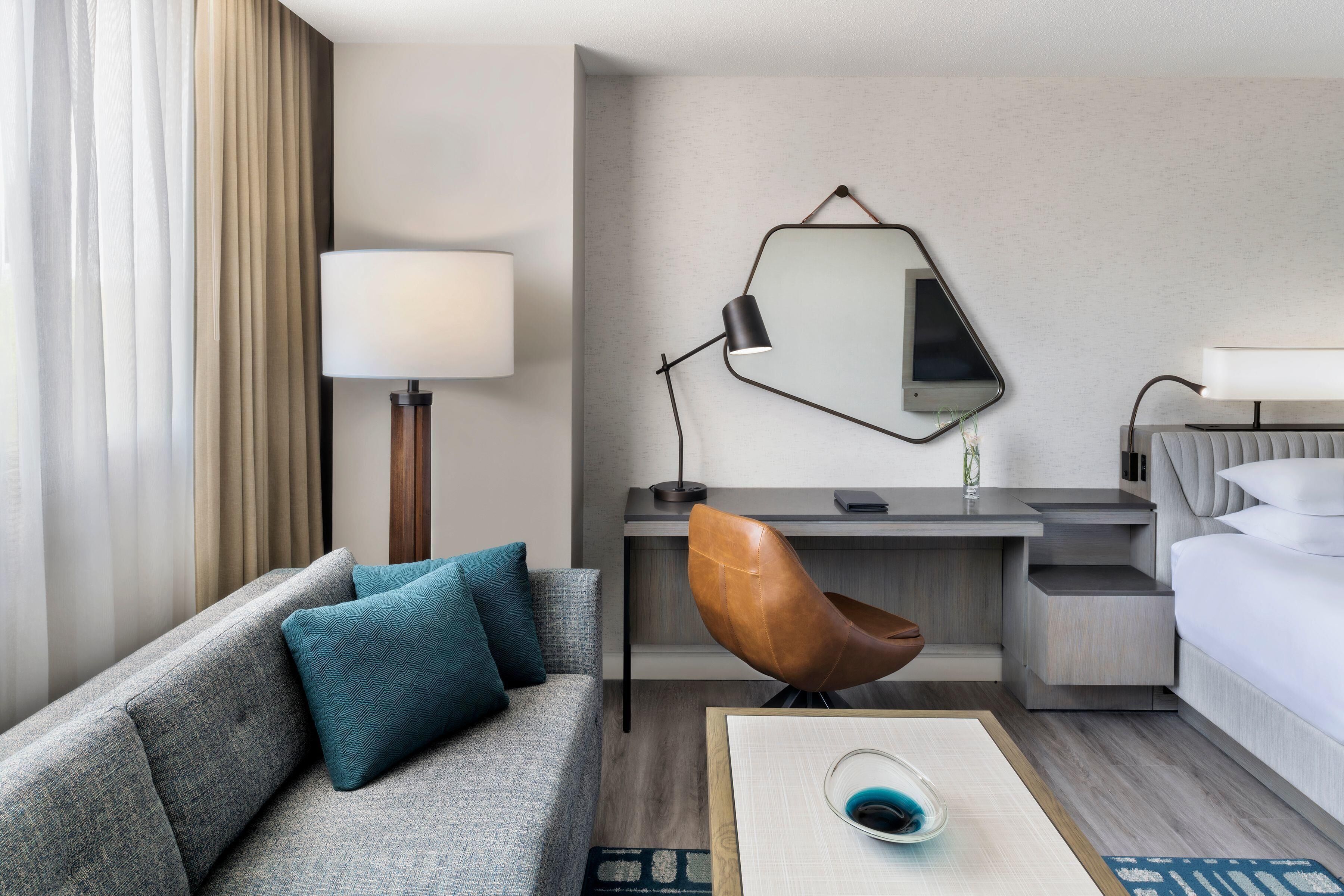 Habitación de hotel con sofá, escritorio, silla, cama, lámpara y espejo.