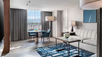 A modern hotel room with a sofa, a dining table with chairs, a TV, a lamp, and a large window with curtains.