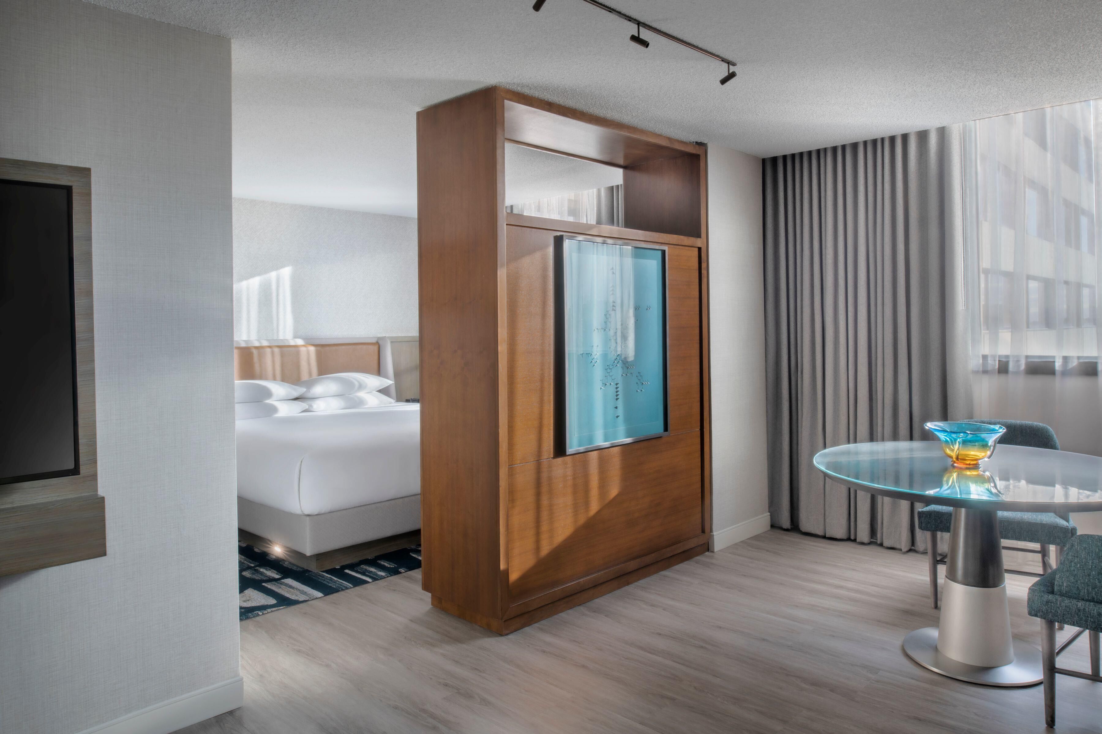 Ein Hotelzimmer mit einem Bett, einem Holzschrank, einem Glastisch mit Stuhl und einem an der Wand befestigten Fernseher.