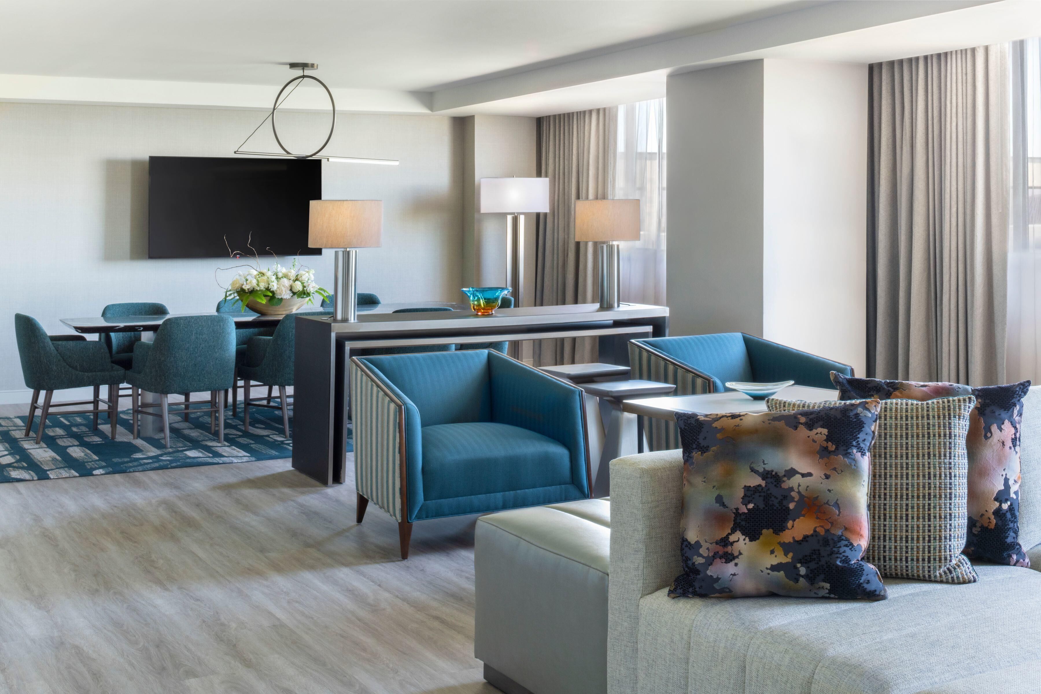 A modern hotel room with a flat-screen TV, a dining area with chairs, a sofa with decorative pillows, and a large window with curtains.