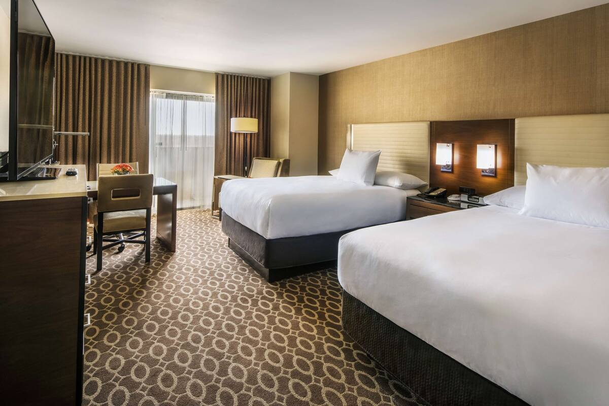 A hotel room with two beds, a desk, a chair, a lamp, and a window with curtains.