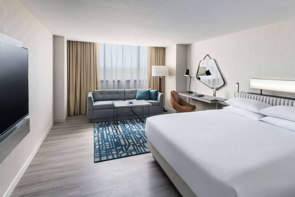 A hotel room with a large bed, a sofa, a TV, a desk with a lamp, and a window with curtains.