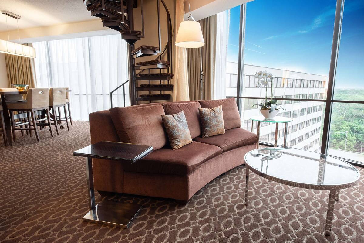 A modern hotel room with a brown sofa, a glass coffee table, a laptop on the sofa, a dining area with chairs and a table, and a large window with a view of a building and greenery.