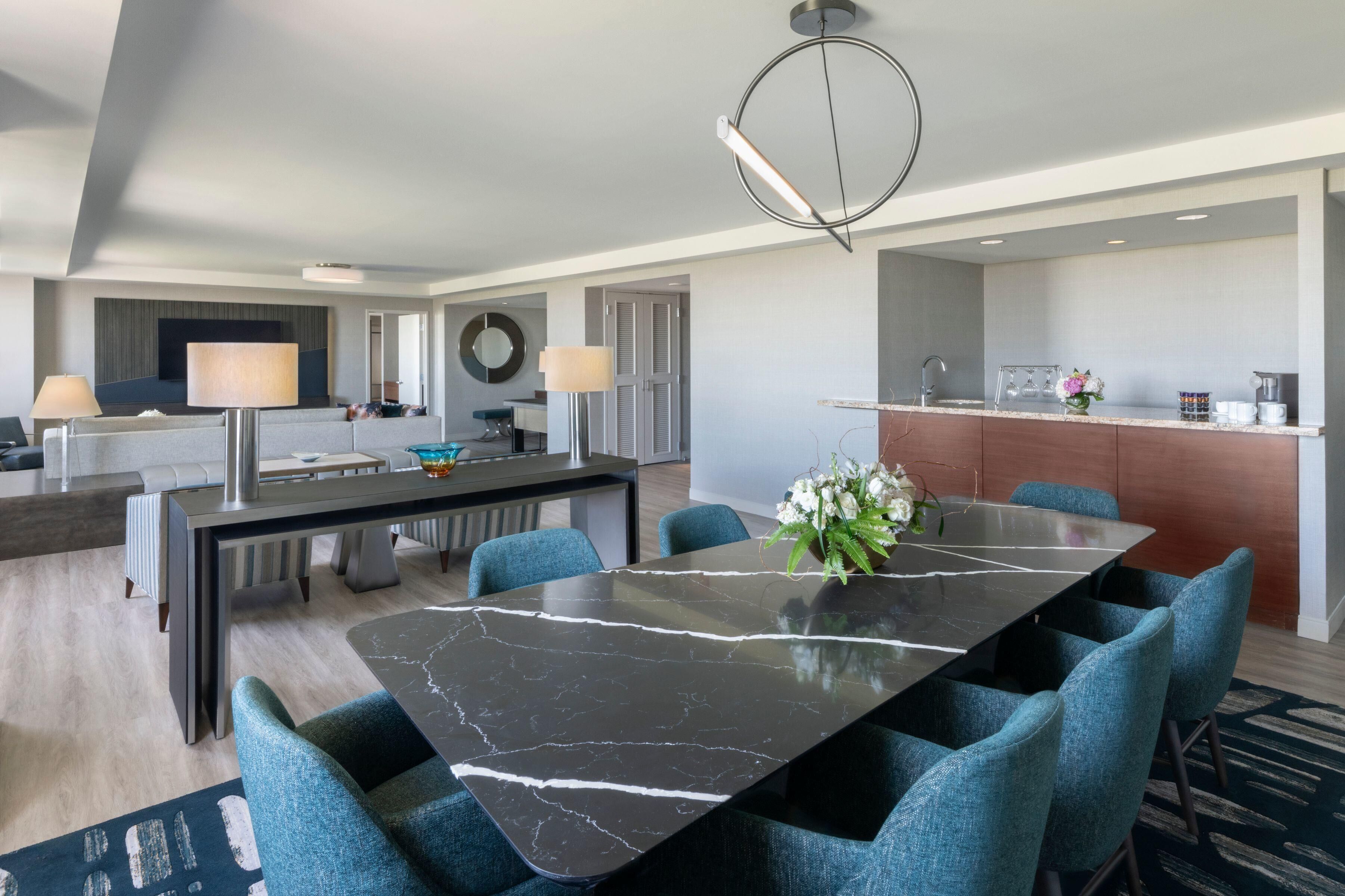 A modern hotel room with a dining area featuring a marble table and blue upholstered chairs, a kitchen with a coffee machine, and a large mirror on the wall.