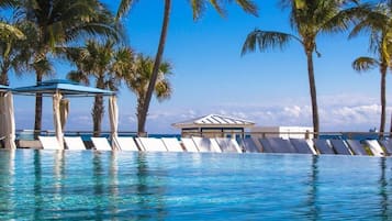 Une piscine d’un bleu éclatant, dotée de chaises longues et d’un pavillon, le tout entouré de palmiers et surplombé par un ciel dégagé.