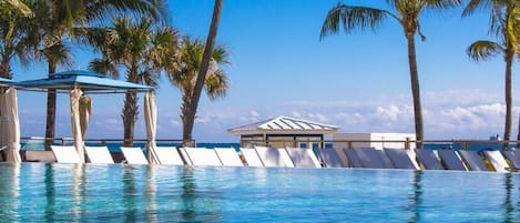 Una piscina azul cristalina con sillas de descanso y un pabellón, rodeada de palmeras y un cielo despejado.