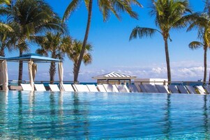 Une piscine d’un bleu éclatant, dotée de chaises longues et d’un pavillon, le tout entouré de palmiers et surplombé par un ciel dégagé.